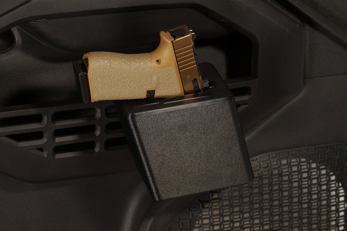 A black plastic gun rack mounted on a dark vehicle interior door panel, holding a tan semi-automatic handgun.
